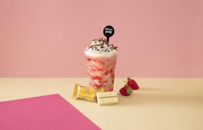 Foto em tons de cor de rosa da receita de smoothie de morango servida sobre uma mesa amarela com kit kat amarelo à frente