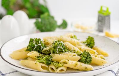 fotografia em tons de branco, bege e verde vista de frente. Contém um prato redondo branco com macarrão tipo penne e brócolis