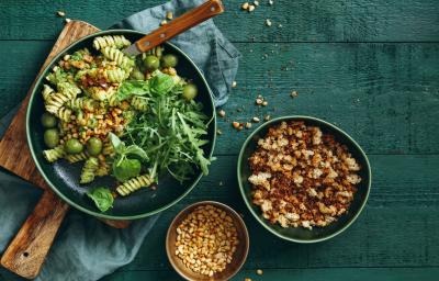 Fotografia vista de cima de uma salada verde com macarrão parafuso com iogurte, azeitona e nozes sobre uma tábua de madeira quadrada. Ao lado, dois bowls com grãos e croutons. A tábua está apoiada em um pano azul, que está sobre uma mesa em tom verde.