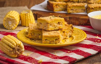 Foto de uma bancada de madeira com uma toalha vermelha quadriculada, com um prato amarelo no centro e pedaços da torta de milho com frango. Há milhos decorando a cena e, ao fundo, mais pedaços de torta sobre uma tábua de madeira.