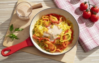 Fotografia em tons claros vista de cima de uma frigideira vermelha e branca com uma receita de moqueca e novasource. A frigideira está sobre uma tábau de madeira retangular grande ao lado de um pano brano com listras vermelhas e tomates pequenos. Fotografia em tons claros vista de cima de uma frigideira vermelha e branca com uma receita de moqueca e novasource. A frigideira está sobre uma tábau de madeira retangular grande ao lado de um pano brano com listras vermelhas e tomates pequenos.