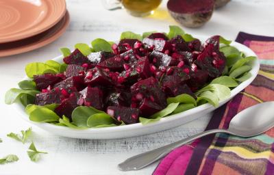 Fotografia em tons de roxo em uma bancada de madeira branca com um pano colorido xadrez com roxo, rosa e amarelo. Ao centro, um recipiente branco oval com salada de beterraba temperada com salsinha e cebola. Ao fundo, beterraba cortada ao meio.