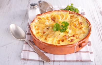 fotografia em tons de branco, vermelho e amarelo de uma bancada branca vista de cima. Contém um recipiente redondo vermelho com um gratinado de batatas, e ao lado uma colher para servir.