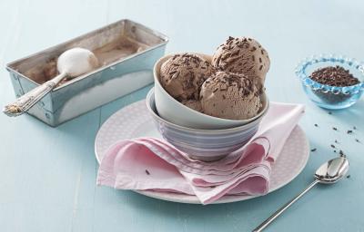 Fotografia em tons de azul e rosa em uma bancada de madeira azul clara, um pano rosa, um pratinho rosa redondo com potinhos de porcelana em cima e as bolas de sorvete de brigadeiro com granulado para decorar. Ao lado, um potinho com granulados e colher.