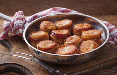 Fotografia em tons de marrom, prata e vermelho de uma bancada de madeira e uma tábua de madeira, sobre ela um panela de inox com medalhões de frango com bacon. Ao lado um garfo de servir, ao fundo um paninho branco e vermelho.