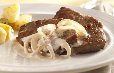 foto em tons de branco e marrom de uma bancada vista de cima. Contém um prato branco redondo com um bife com cebolas com creme e abaixo um pano branco.