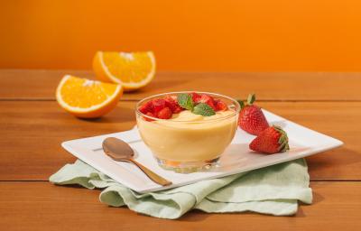 Fotografia em tons de laranja em uma bancada de madeira, um fundo laranja, um recipiente branco retangular, um pano verde claro e uma tacinha de vidro com a mousse de laranja dentro dela, com morangos para decorar. Ao fundo, uma laranja partida ao meio.