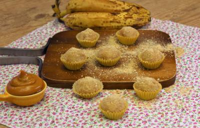 Fotografia em tons de amarelo em uma bancada de madeira de cor marrom. Ao centro, uma toalha branca florida e em cima, há uma tábua de madeira contendo os brigadeiros. Ao fundo, há um cacho de bananas.