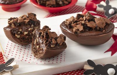 Fotografia em tons de branco e vermelho de uma bancada branca com uma tábua branca e uma paninho branco com bolinhas vermelhas, sobre ela dois ovos de pascoa recheados. Ao lado coelinhos. Ao fundo dois recipientes vermelhos com brownie e chocolate.