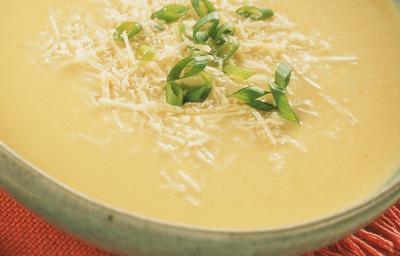 Fotografia em tons de creme e vermelho, com refratário cinza ao centro contendo um creme amarelo decorado com queijo e cebolinha picada sobre guardanapo vermelho, com colher ao lado e tudo sobre aparador de palha na cor bege.