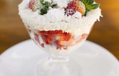 Fotografia de uma taça de sobremesa com merengue, morangos, suspiro e granulado branco nas bordas. O recipiente está em cima de um prato branco de sobremesa, com uma colher pequena. O prato está sobre uma mesa marrom.