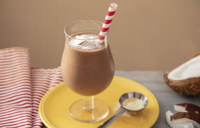 Fotografia em tons de marrom em uma bancada de madeira de cor cinza. Ao centro, um prato amarelo contendo uma taça com o smoothie. Ao fundo, há um pano branco listrado de vermelho e um coco partido ao meio.