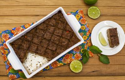 Fotografia em tons de marrom em uma mesa de madeira com um pano florido, um refratário com alças laterais brancas com o quibe de carne dentro dele. Ao lado, um pratinho branco com uma fatia do kibe e rodelas de limão.