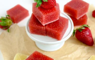 Fotografia em tons de vermelho em uma bancada de madeira com um pano bege, um suporto de bolo branco, com balas de goma de morango. Ao lado, morangos e fatias de limão siciliano.