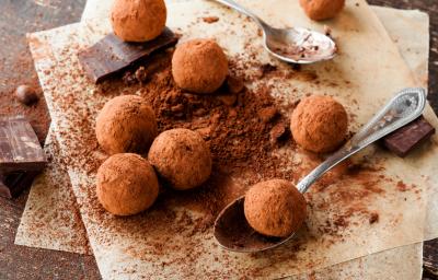 Fotografia vista de cima de algumas trufas de café e chocolate passadas no cacau em pó, sobre uma tábua de madeira salpicada com cacau em pó. Tem alguns pedaços de chocolate em volta das trufas e uma colher segurando uma delas.