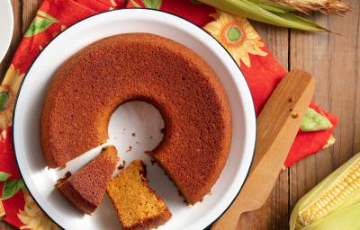 Bolo de milho feito com Farinha Láctea Nestlé em um prato branco, com dois pedaços cortados e espigas de milho em volta.