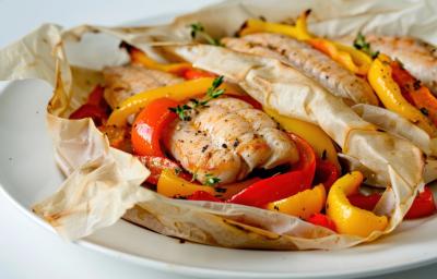 Fotografia de um filé de peixe no papillote com legumes por baixo cortados em fatias. Na fotografia são dois papillotes de peixe com legumes, um do lado do outro sobre um prato branco de vidro.