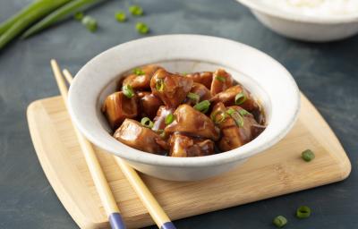 Fotografia em tons de marrom em uma bancada de madeira cinza escura com uma tábua de madeira ao centro, com um prato redondo fundo bege e o frango teriyaki com cebolinhas verdes dentro dele. Ao lado, hashis apoiados na tábua.