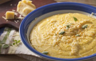 Fotografia em tons de marrom e azul de uma bancada marrom com um paninho branco, sobre ele um recipiente redondo branco e azul com polenta. Ao fundo uma tábua com queijo parmesão.