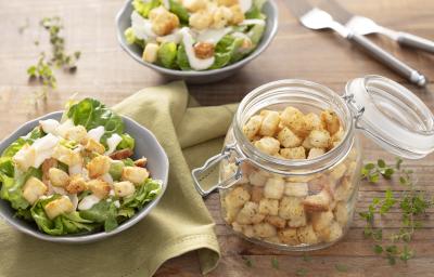 Fotografia em tons de verde em uma bancada de madeira clara, um pano verde, dois bowl com os croutons com a salada de alface e molho de iogurte. Ao centro, um pote de vidro hermético com vários croutons dentro dele.