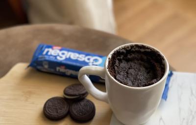 Foto de uma caneca branca contendo a receita de Bolo Negresco, sobre uma tábua com alguns Biscoitos Negresco soltos e um pacote em tom azul de Biscoito Negresco