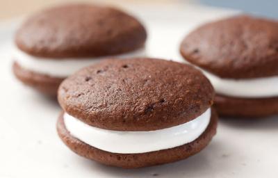 Fotografia em tons de preto em uma bancada de madeira clara, um prato redondo branco raso com três sanduicinhos de chocolate recheados com marshmallow.