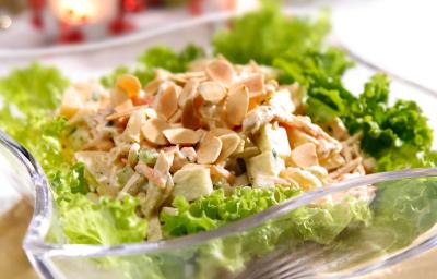 Fotografia em tons de verde em uma mesa com uma toalha branca, um apoiador de prato de madeira, um recipiente de vidro fundo com a salada de alface e frango dentro dele.