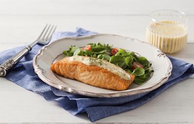 fotografia em tons de branco e azul de uma bancada branca vista de frente, contém um pano azul, um prato redondo branco com um pedaço de salmão com creme branco por cima e folhas com fatias de tomate cereja para acompanhar. Ao lado um garfo para servir