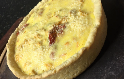 Foto em tons de amarelo da receita de quiche de frango com tomates secos servida em uma porção sobre uma mesa de pedra preta bem escura