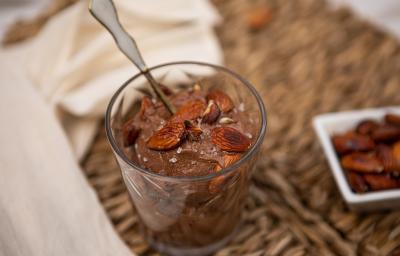 Fotografia de uma mistura de chocolate meio amargo, nozes e creme de leite em um recipiente de vidro. Por cima, nozes, flor de sal e uma colher sobre o creme. Ao lado, um pano branco e um recipiente branco de vidro com nozes.