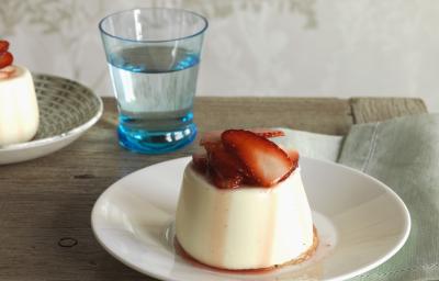 Fotografia de um flan de canela com calda de morangos em um prato branco fundo sobre uma mesa de madeira. Ao fundo, outro flan de canela e, ao lado, um copo d'água.