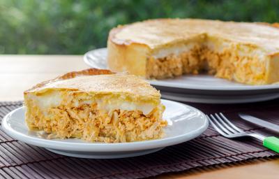 Fotografia de uma fatia de torta de frango com requeijão sobre um prato pequeno de vidro raso. No fundo tem um apoio branco e preto com o restante da torta quase inteira.