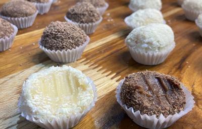 Fotografia em tons de marrom e branco em uma bancada de madeira de cor marrom. Ao centro, estão várias forminhas enfileiradas com brigadeiros de chocolate e brigadeiro branco.