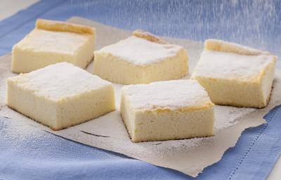 Fotografia em tons de amarelo, azul e branco de uma bancada branca com paninho azul, sobre ele pedaços de torta de ricota polvilhadas com açúcar.