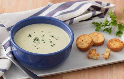 Fotografia em tons de azul e verde, com cumbuca azul ao centro com creme de legumes e salsa picada com torradinhas sobre travessa branca, ramo de salsa, brócolis e couve-flor decorando, guardanapo branco e azul com colher, sobre bancada de cor palha.