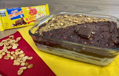 Fotografia em tons de amarelo e vermelho com uma travessa de sobremesa ao centro. Na travessa existe uma sobremesa a base de chocolate e coberta com pedações de amendoim e tabletes de chocolate ao leite. Ao fundo existe uma barra de chocolate fechada.