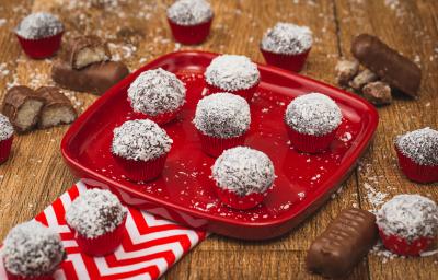 Imagem de uma prato vermelho com algumas unidades de Brigadeiro marrom passado em flocos de coco. A bancada está decorada com pedaços de bombons, um tecido vermelho e branco e mais brigadeiros.