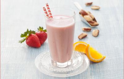 Fotografia em tons de rosa em uma bancada de madeira cinza com uma taça de vidro com a vitamina de morango com laranja dentro dele. Ao fundo, morangos, laranja e castanhas espalhados pela mesa.