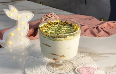 Fotografia de uma taça de vidro servido com camadas de creme, biscoito e pasta de pistache. Ao fundo, um pano rosa e uma luminária em formato de coelho.