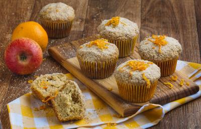 Imagem aproximada de muffins em tom claro, sobre uma tábua de madeira em um pano quadriculado em tom amarelo e branco. Ao fundo há uma maçã e uma laranja e alguns farelos de Farinha Láctea.