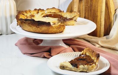 Fotografia em tons de marrom em uma bancada de mármore com um pano marrom em volta e um prato branco elevado com a torta fácil de café ao centro e ao lado um pratinho com um pedaço  da torta. Ao fundo, uma tábua de madeira.