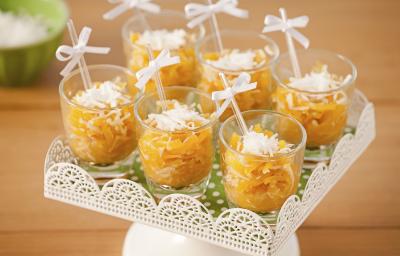 Fotografia em tons de laranja em uma mesa de madeira com um suporte de doces branco ao centro com os doces de abóbora em copinhos transparentes com palitinhos e um lacinho branco.