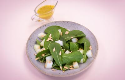 Fotografia em tons de rosa e verde em uma mesa de madeira rosa com um prato redondo raso lilás e a salada de rúcula com limão dentro dele. Ao fundo, um potinho com azeite.