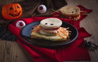 Imagem aproximada da receita de um sanduíche em formato de aranha, com pão, patas feitas com tiras de cenoura, além de o cenário estar decorado com itens de Halloween