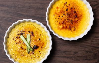 Fotografia vista de cima de dois ramekins brancos, um com creme brulée de baunilha com laranja, e o outro creme brulée de aspargos com bacon. Os ramekins estão sobre uma mesa de madeira.