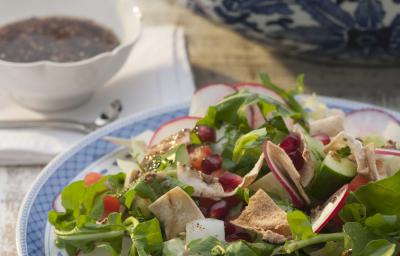 Fotografia em tons de verde em uma bancada de madeira com um prato branco com detalhes em azul e a salada com agrião, rabanete, alface dentro. Ao fundo, um potinho com molhinho.