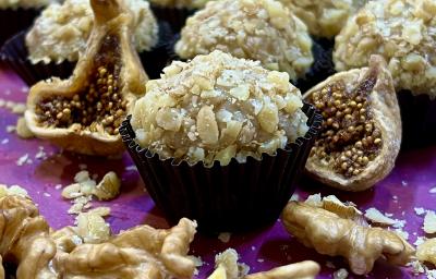 Foto de receita de Brigadeiro de Figo com Nozes. Observa-se o brigadeiro com nozes picadas em volta em uma forminha preta, com figos secos cortados ao lado e nozes na projeção da frente. Ao fundo, mais brigadeiros.