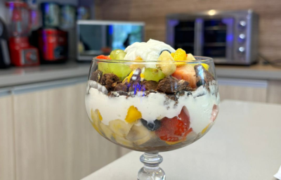 Fotografia de uma taça grande de vidro com pavê de frutas e iogurte, O pavê está sobre uma bancada e no fundo tem aparelhos eletrodomésticos.