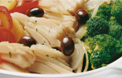 Fotografia em tons de verde e vermelho em uma bancada de madeira em uma mesa com uma toalha cinza, um refratário oval branco com bacalhau com tomate, batata e brócolis.