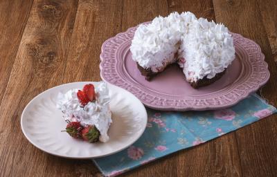 Imagem da receita de Torta Brownie, decorada com chantilly e morangos, servida em um prato rosa e ao lado há um prato menor branco com uma fatia. Tudo está sobre um pano decorado com motivos florais, sobre uma bancada de madeira